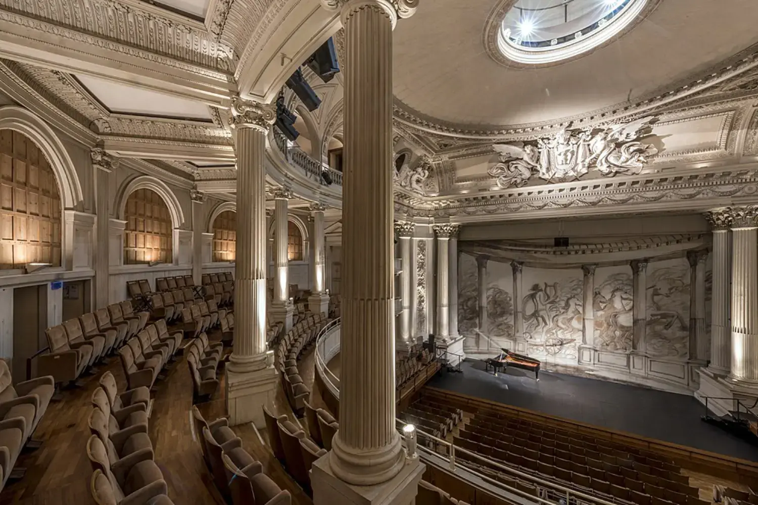 Elégant théâtre historique de l'Hôtel à Compiègne avec des colonnes ornées, un plafond détaillé et des gradins vides faisant face à une scène avec un piano et des peintures murales.