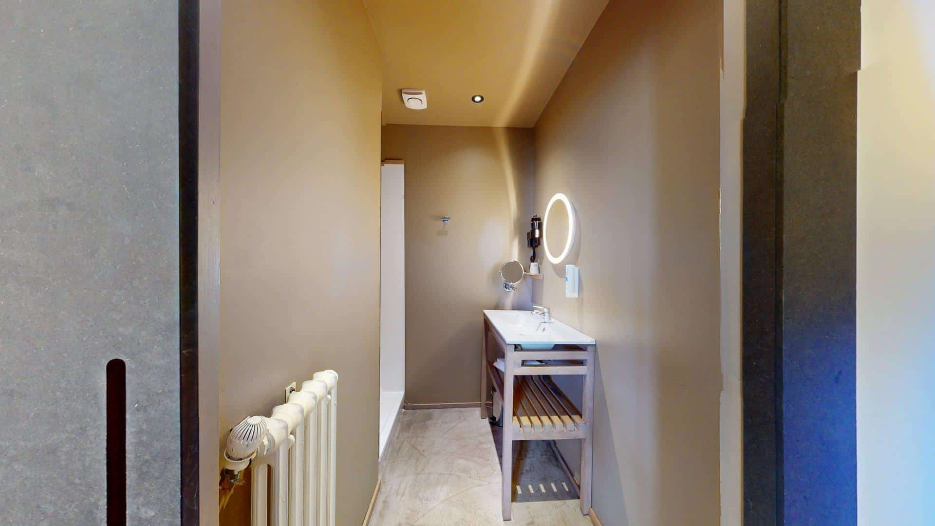 Salle de bain moderne à l'Hôtel de Harlay avec des murs beiges, un lavabo blanc sur un support en bois, un miroir rond éclairé, un radiateur et une douche à l'italienne - parfait pour votre séjour dans notre Hébergement Compiègne.