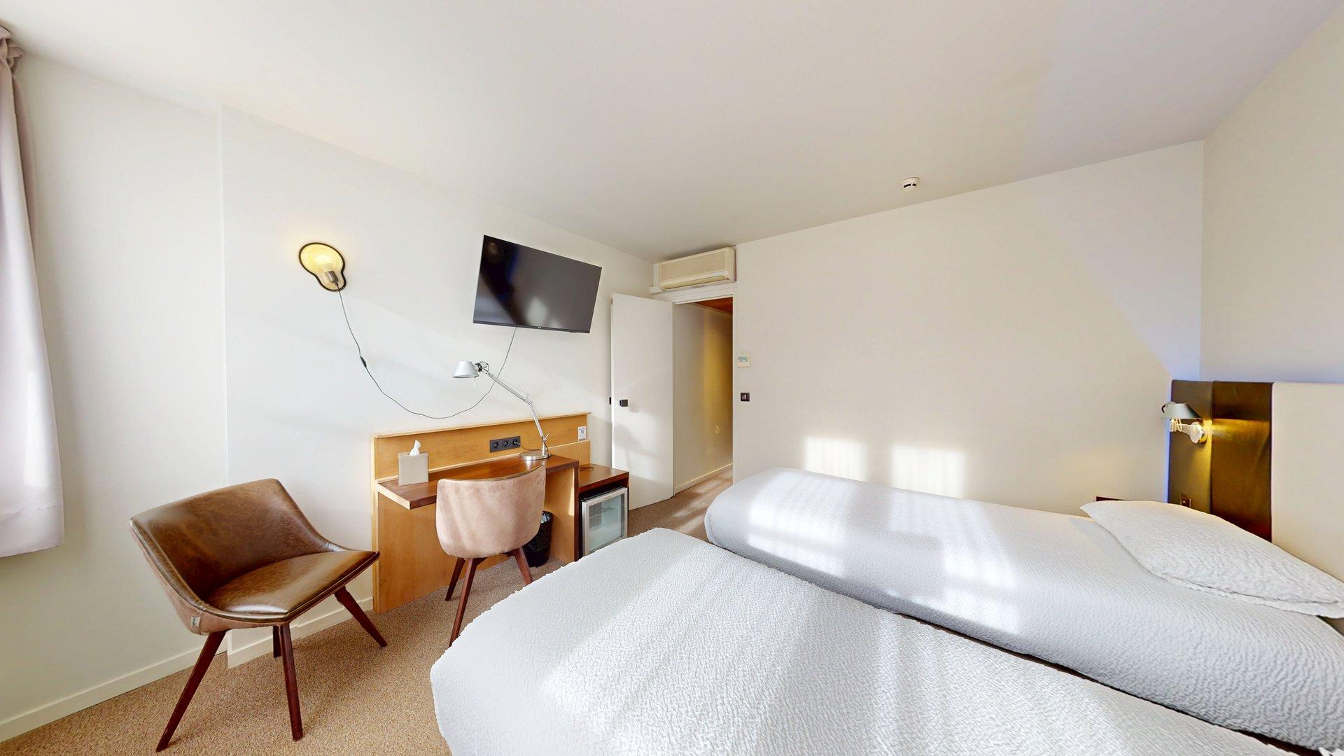 Chambre d'hôtel à l'Hôtel de Harlay avec deux lits simples, un bureau et une chaise, une télévision murale, un petit réfrigérateur et un décor neutre. La lumière du soleil pénètre par une fenêtre sur la gauche - idéal pour un hébergement confortable à Compiègne.