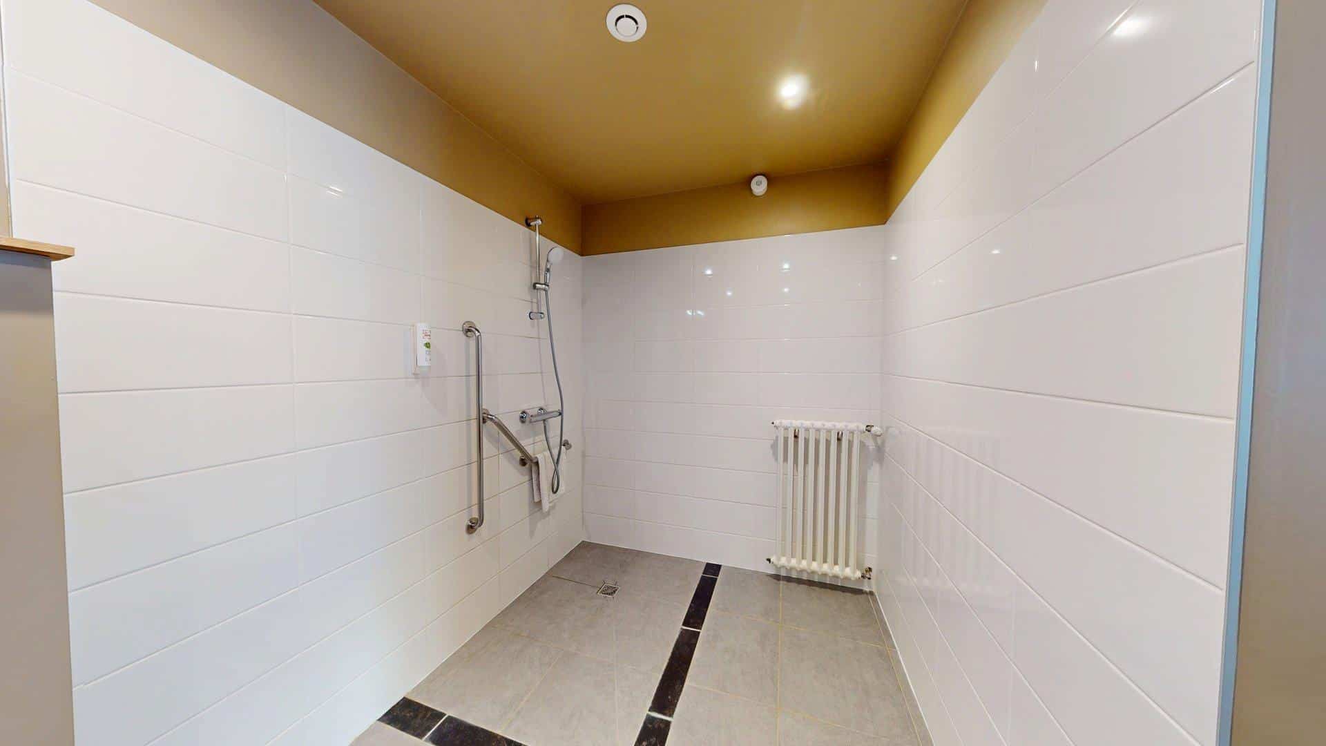 Salle d'eau accessible à l'Hôtel de Harlay avec douchette, barres d'appui, murs carrelés blancs, radiateur et plafonniers - assurant un hébergement confortable à Compiègne pour tous les clients.