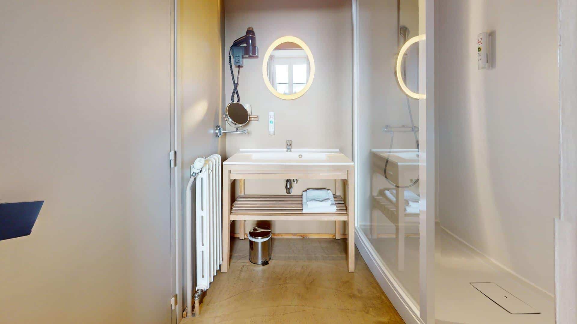 Modern bathroom at Hôtel de Harlay with a wall-mounted sink, round mirror with light, open shelf with towels, hairdryer, trash bin, radiator, and a glass shower enclosure—ideal for your Hébergement Compiègne stay.