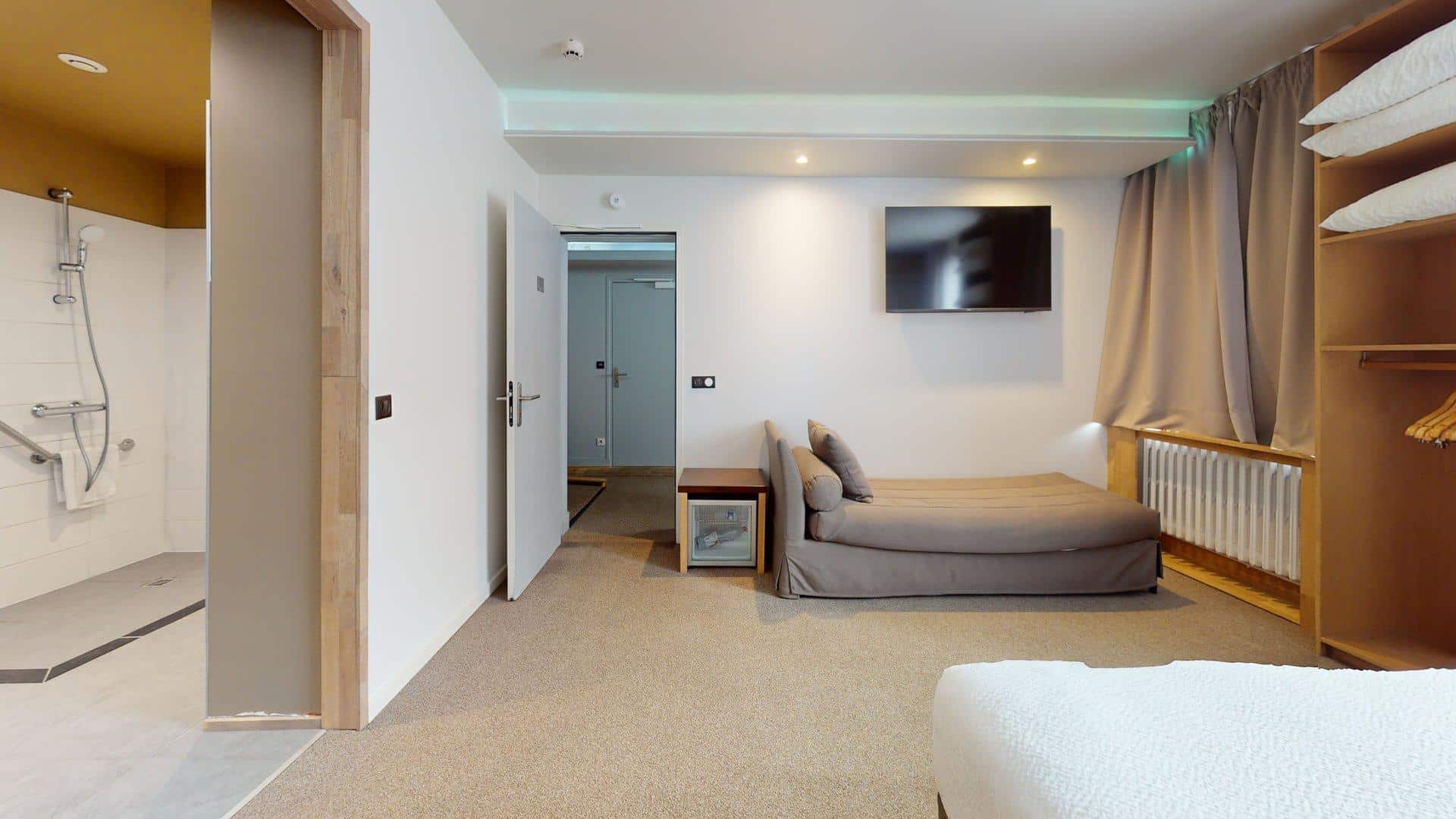 Chambre d'hôtel moderne à l'Hôtel de Harlay, Hébergement Compiègne, avec une salle de bain accessible, un canapé-lit simple, une télévision murale, une armoire ouverte et un décor neutre.